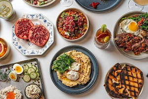 A spread of dishes: grilled meat, bowls with chopped toppings, sunny-side eggs, a parmesan crusted pie, cucumber salad, hash browns, and herbs, all colorful and plated.