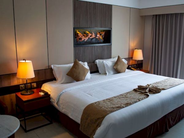 A modern hotel room with a large bed, decorative pillows, and warm lighting. A faux fireplace adds ambiance above the bed.