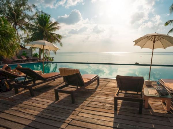Lounge chairs and umbrellas are on a wooden deck by a calm infinity pool, with palm trees and a serene ocean view in the distance.
