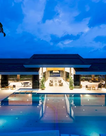 A luxurious modern building with a large swimming pool, lit up beautifully against a cloudy evening sky.