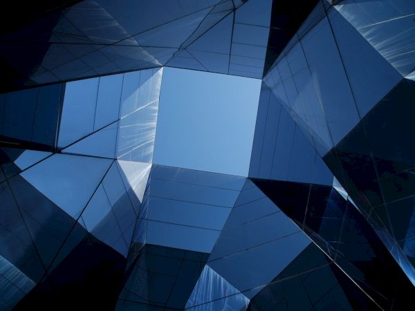 The image shows an architectural view upwards through dark blue glass panels forming geometric shapes with a central opening to the sky.