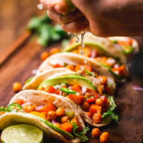 The image shows tacos filled with veggies and chickpeas, garnished with avocado slices and cilantro, with lime being squeezed on top.