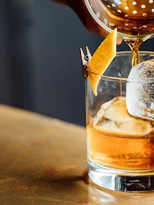 A drink is being poured into a glass with ice and a garnish on a wooden surface.