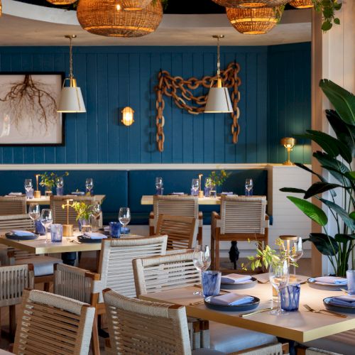 A stylish seaside restaurant with blue walls, wicker chairs, plank tables set with blue napkins and glassware, hanging lights, and leafy plants, creating a relaxed coastal vibe.