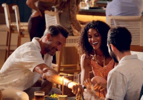 Three friends enjoy a cozy rooftop dinner, sharing drinks and laughter around a small table under warm string lights.