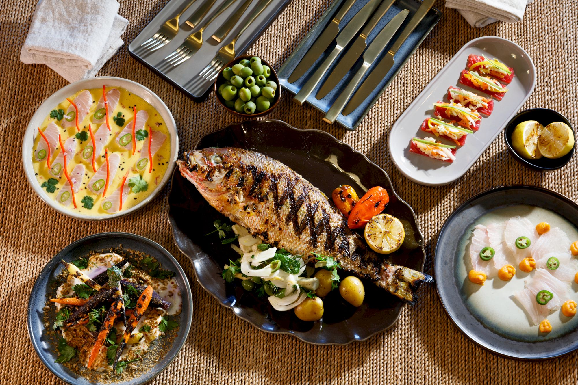 A feast spreads: a roasted fish centerpiece with lemon, sides of creamy soup, a herb salad, grilled veggies, a baked dish, and plated bites for sharing.