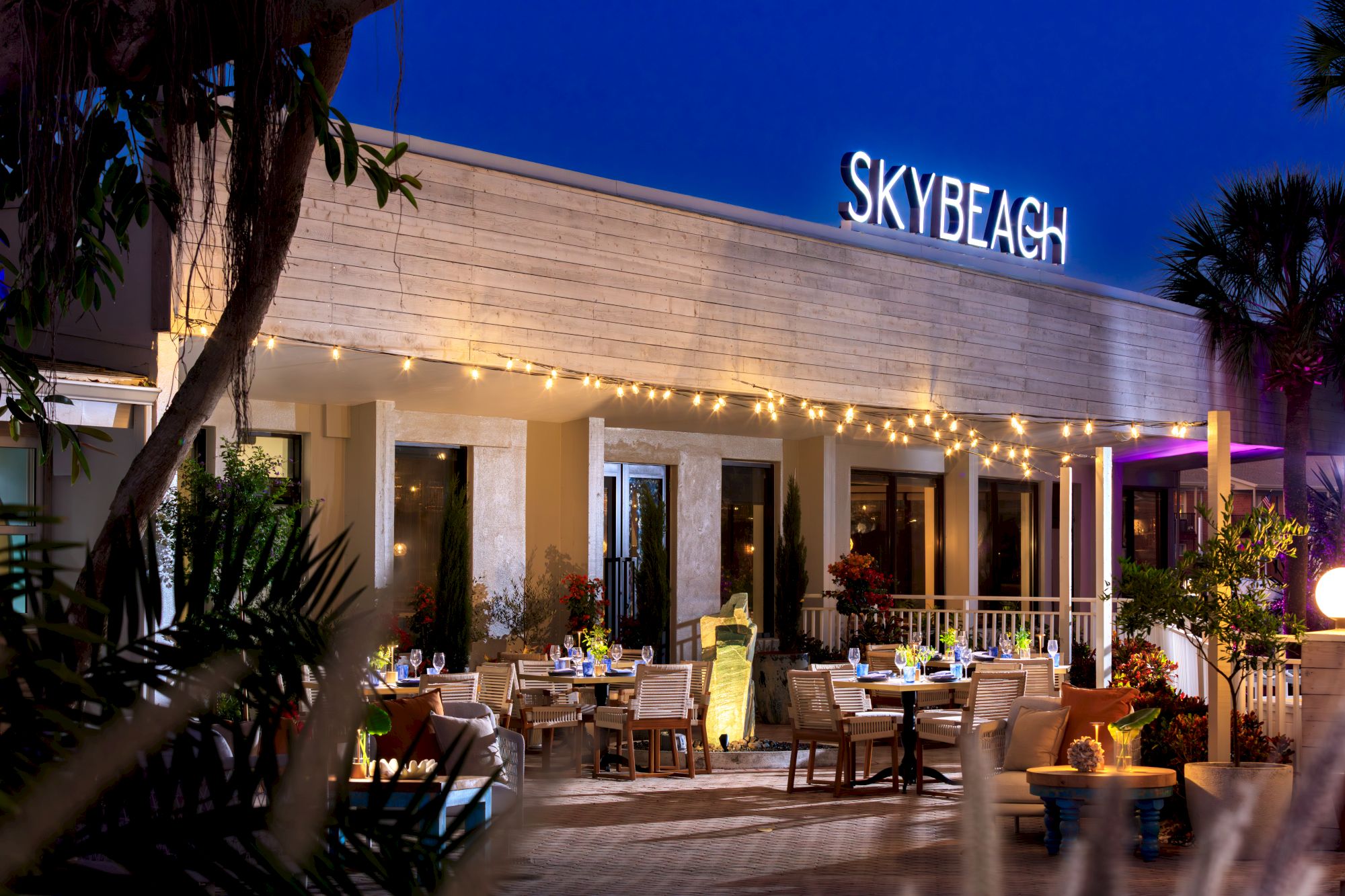 A stylish outdoor restaurant called Sky Beach at dusk, with string lights, cozy tables, and tropical plants by the entrance.
