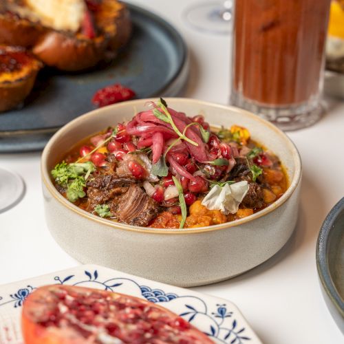 A colorful bowl of grain or rice topped with herbs, pickled onions, vegetables, and meat; surrounded by bread, fruit, and a tall iced tea.
