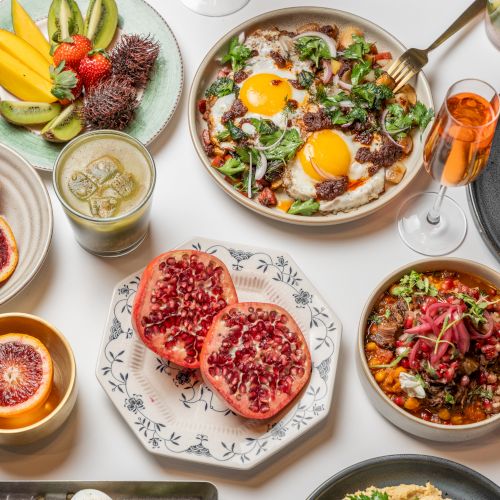 A colorful brunch spread: eggs sunny-side up with greens, vibrant fruit, pita chips, bowls of grains and veggies, pomegranate, juices, and pastries, all on a bright table.