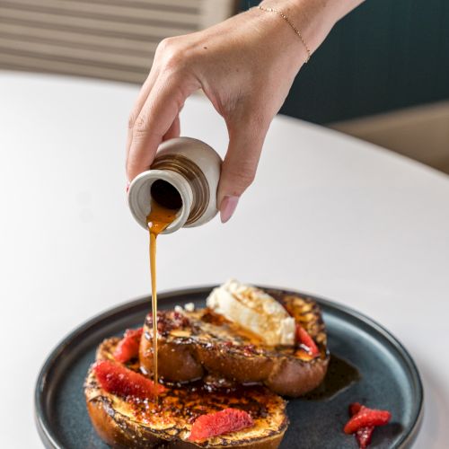 A hand pours syrup over a plate of bread topped with fruit and cream, drizzling caramel-like liquid onto the dessert plate. ending.