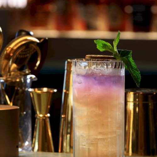 A tall colorful cocktail with mint garnish on a bar counter, set against a glow of gold barware in the background.