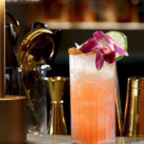 A pink cocktail garnished with a flower on a bar counter, with gold barware in the blurred background.