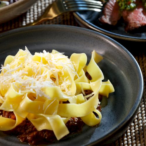 Creamy pasta with shredded cheese over a rich tomato sauce, plated on a dark dish with another meat dish in the background.