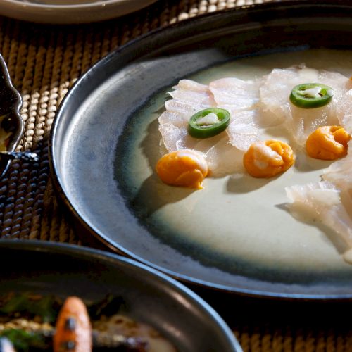 Creamy white fish in milk sauce, topped with sliced green chilies and orange dollops, served on a blue plate with other dishes nearby.