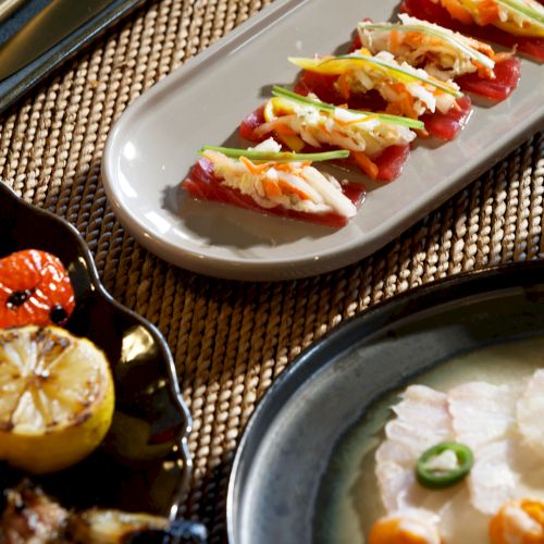 Assorted bite-sized appetizers on a long plate with colorful toppings and greens, plus other dishes on the table.