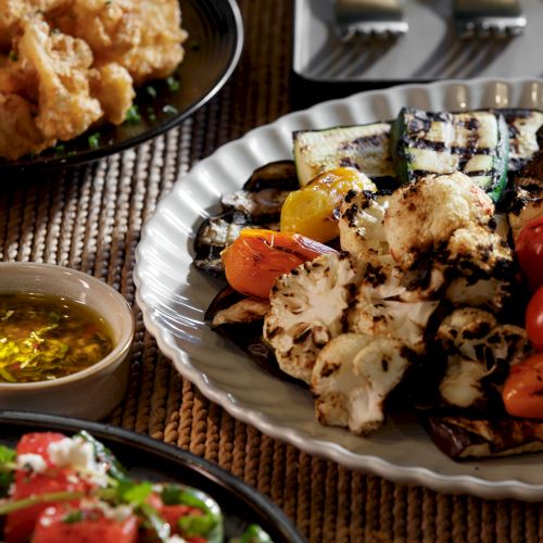 A plate of grilled cauliflower with herbs, roasted peppers, zucchini, and tomatoes sits beside a small bowl of herbed oil, plus other dishes around.