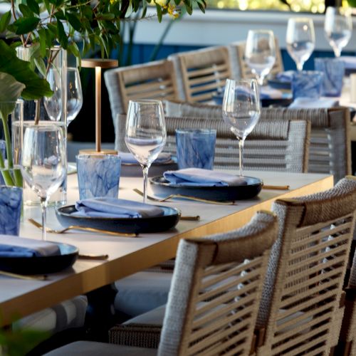 A tidy restaurant table setting with wine glasses, plates, blue napkins, and woven chairs ready for guests, outdoors.