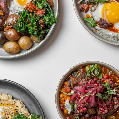 A top-down view of four bowls with hearty bowls of meat, veggies, eggs, potatoes, greens, and colorful toppings, all plated stylishly.