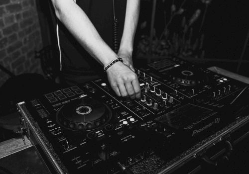 A DJ is mixing on a Pioneer turntable setup, hands on the mixer, dark club vibes, black-and-white photo.