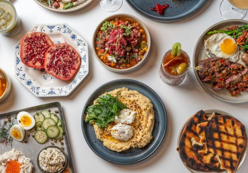 A spread of dishes: steaks, bowls, fried eggs, grilled meat, mashed potatoes with herbs, grilled flatbread, and sides on blue/white ceramics.