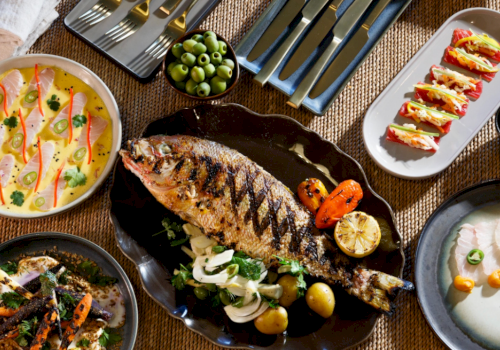 A lavish spread: grilled fish centerpiece with herbs, creamy sauces, quiches, olives, vibrant veggies, small bites, and assorted sides for sharing.