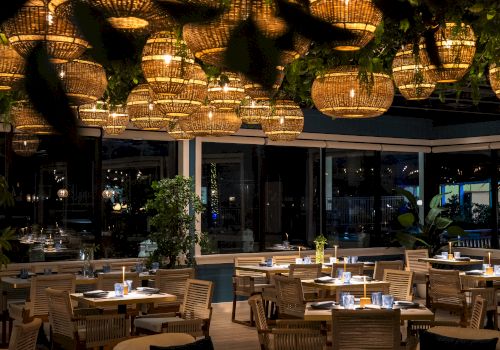 A stylish restaurant with warm hanging lanterns, wicker chairs, and neatly set tables for dinner, creating a cozy, chic dining atmosphere.
