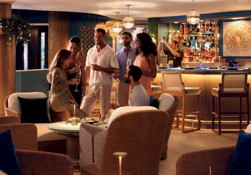 A group of casually dressed people chat and mingle in a cozy lounge with a bar, warm lighting, and modern wicker seating, relaxed party vibes.