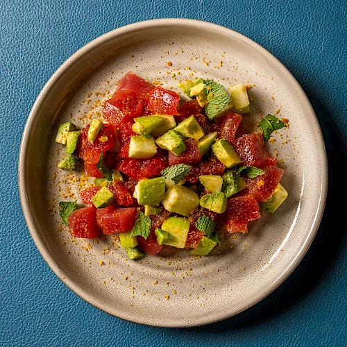 A small plate of diced watermelon and avocado, sprinkled with pepper and lime, set on a blue textured background.