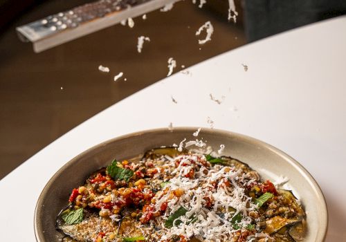 A cook grates white cheese over a hot, saucy eggplant/vegetable dish in a bowl, sprinkling toppings for a finishing touch.