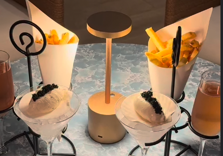 Two martini cocktails with black garnish and yellow chip cones on a bar table by a circular lamp.