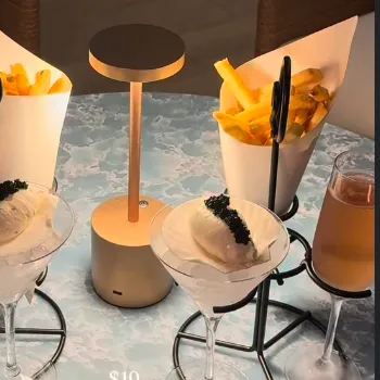 Two martini cocktails with black garnish and yellow chip cones on a bar table by a circular lamp.