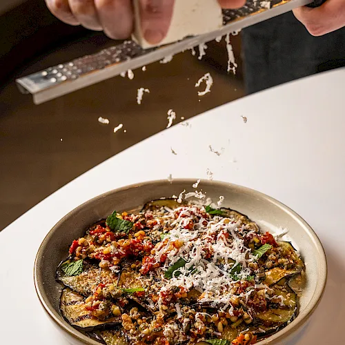 A cook grates white cheese over a hot, saucy eggplant/vegetable dish in a bowl, sprinkling toppings for a finishing touch.