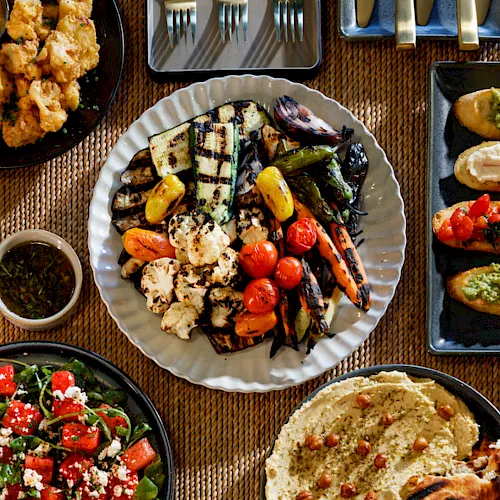 A spread of grilled vegetables, salad, falafel/hummus with pita, and dipping sauce on a table with serving utensils.