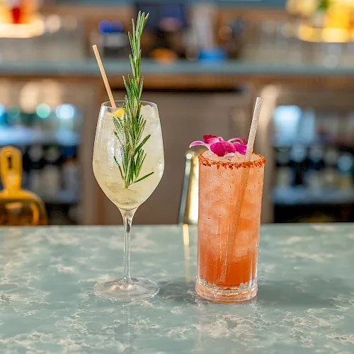 Two cocktails on a bar—one with rosemary garnish and one pink-red drink with ice and a straw.