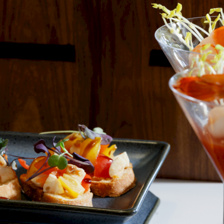 Mini open-faced toast bites topped with colorful vegetables and microgreens, with a glass of sprouts on a wooden table.