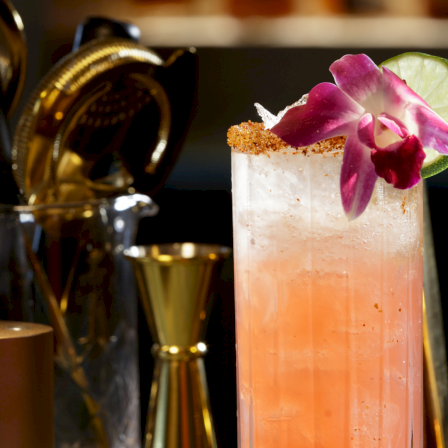 A tall pink cocktail with ice, garnished by a purple orchid and lime slice, with a gold bar setup in the background.