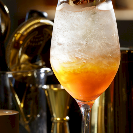 A chilled golden-orange cocktail in a stemmed glass, with bubbles and ice, surrounded by bar bottles.