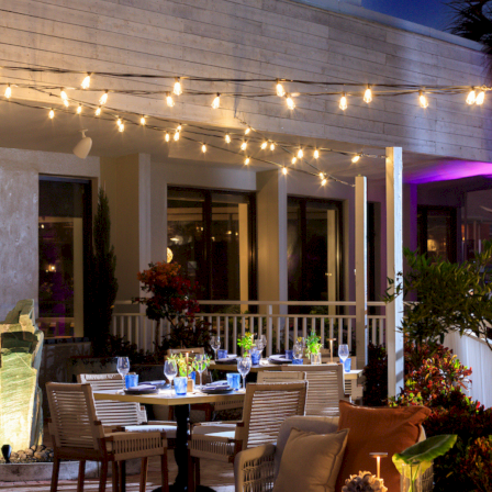 Outdoor patio dining area with a table, chairs, flowers, and warm string lights under a covered walkway.