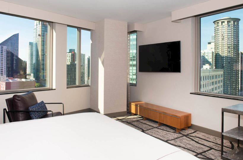 A modern hotel room with a large bed, city skyline views, flat-screen TV, and stylish furniture.