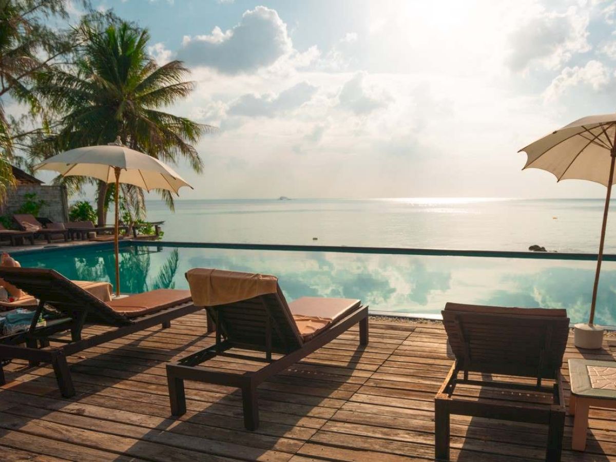 A tranquil beach setting with lounge chairs, umbrellas, and palm trees beside a pool overlooking the ocean under a partly cloudy sky.