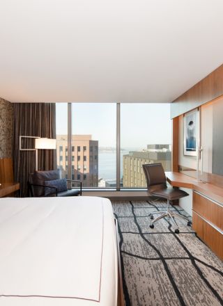 A modern hotel room with a large bed, flat-screen TV, and desk with a city view through the windows, featuring sleek wood accents.