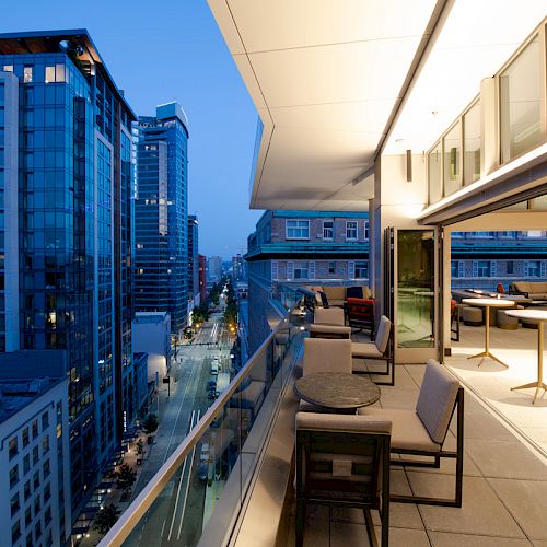 A modern rooftop terrace with seating, overlooking a city street flanked by tall, illuminated buildings under a twilight sky.